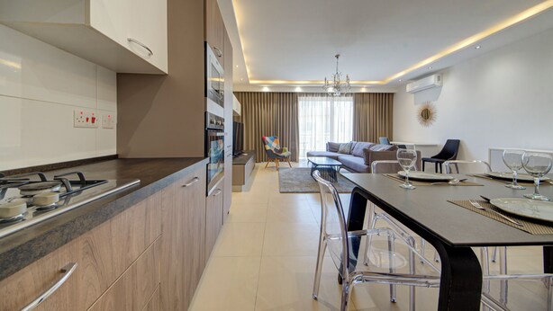 A modern kitchen with a central island, a dining area with a table set for four, and a living room with a sofa and a TV. - Swieqi, Luxury Apartmen/3BD- 2BH,sleeps 6 (Is-Swieqi)