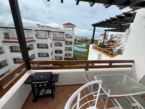 A balcony with a grill, a table, and a chair, overlooking a pool and buildings. - Beachfront Apartment, Private Solarium, 4 Pools, Secure Residence, WiFi, Parking (Asilah)