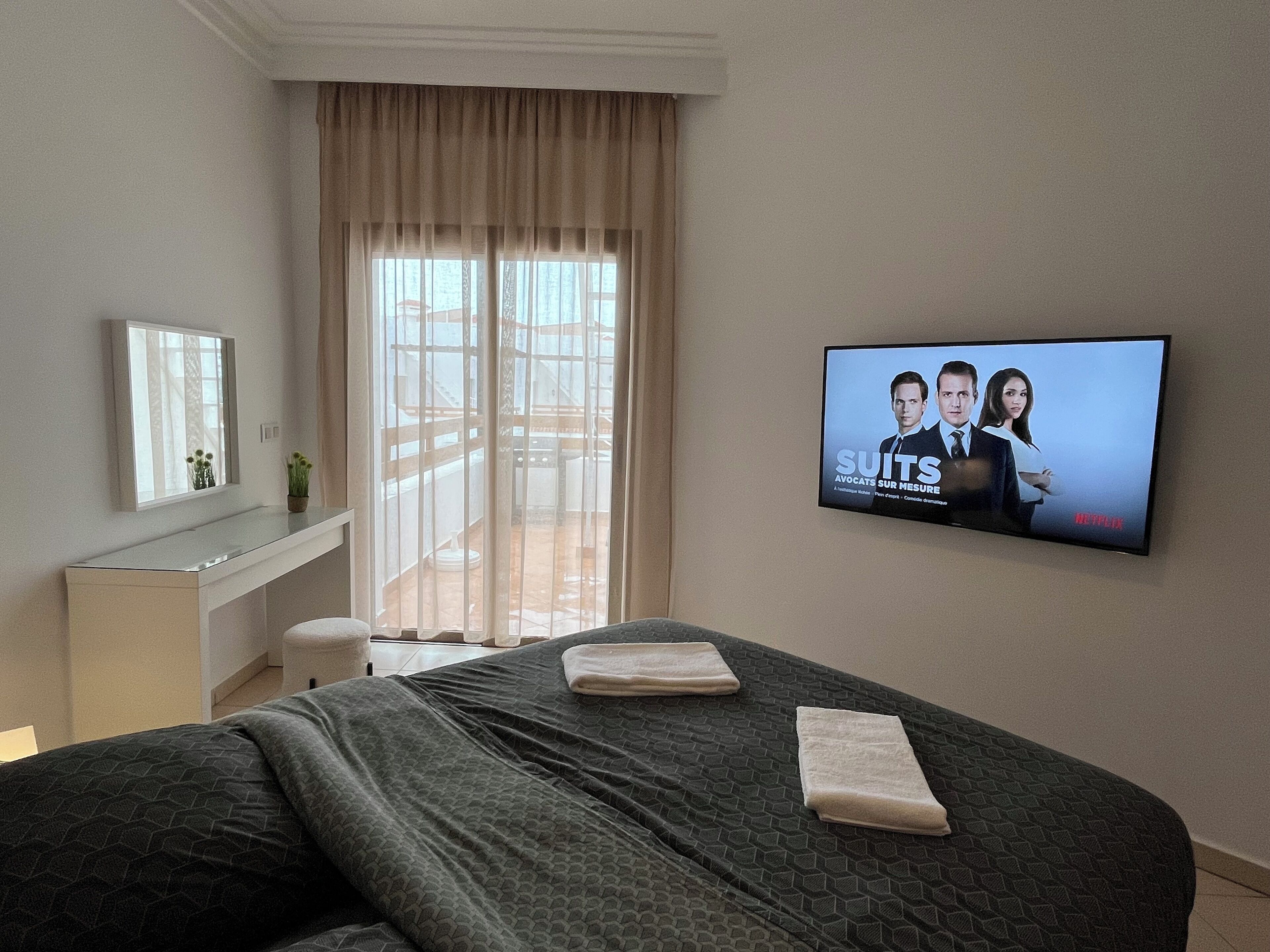 Hotelkamer met een bed, een tv waarop een film draait, een raam met gordijnen en een klein bureau met een plant.