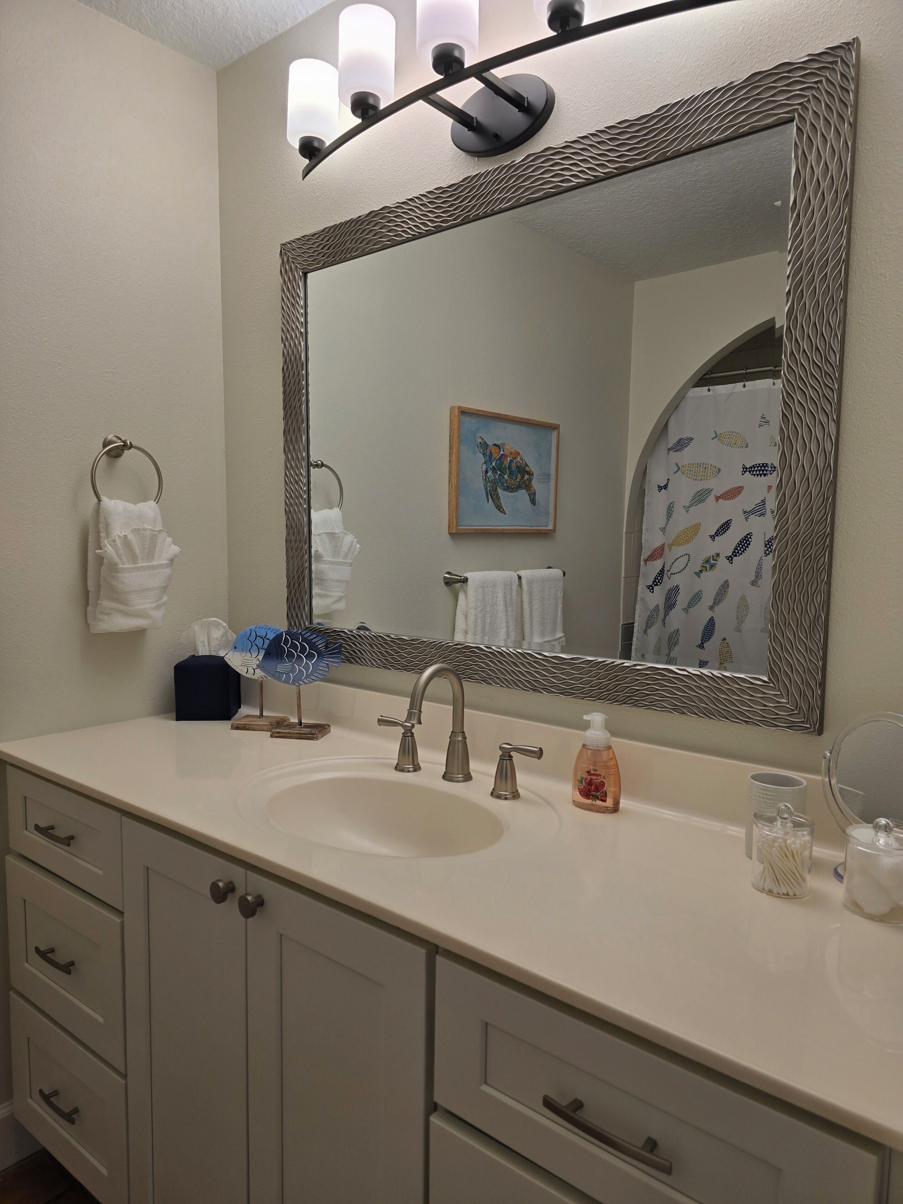 A bathroom with a large mirror, a sink, two faucets, a towel rack, a framed picture, and a shower curtain with fish patterns.