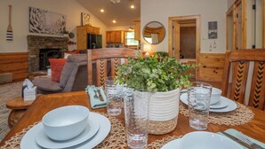 A dining area with a wooden table set for four, surrounded by chairs, a fireplace, and a kitchen in the background. - Riverfront Little Red Trout Cabin, Hot Tub & Views (Heber Springs)