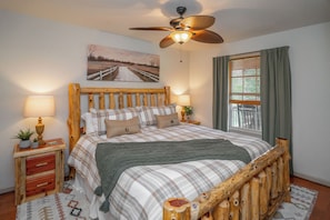 A bedroom with a wooden bed frame, a ceiling fan, a painting on the wall, and a window with curtains.