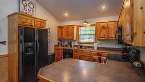 A kitchen with wooden cabinets, a refrigerator, a dishwasher, a coffee maker, and a window with blinds. - Riverfront Little Red Trout Cabin, Hot Tub & Views (Heber Springs)