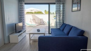 A living room with a sofa, a TV on a stand, and a sliding glass door leading to an outdoor area with a table and chairs. - Riboleys - Villa 10 personnes avec piscine privée à 200m de la mer (Vias)