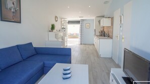 A modern living room with a blue sofa, a white coffee table, and a TV. The room leads to a kitchen with white cabinets and a dining area with a table and chairs. - Riboleys - Villa 10 personnes avec piscine privée à 200m de la mer (Vias)