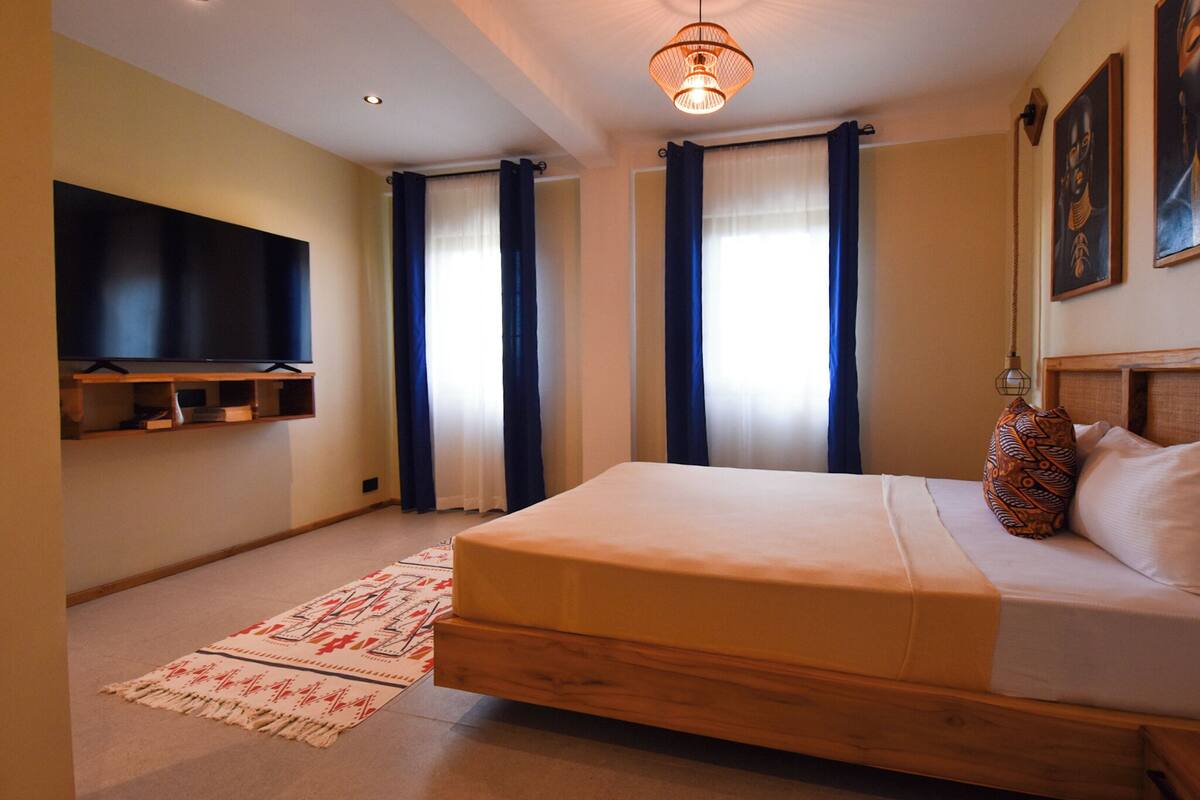 A bedroom with a bed, a flat-screen TV mounted on the wall, and a window with curtains.