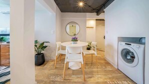 A compact living space with a round dining table, white chairs, a round mirror, a washing machine, and potted plants.