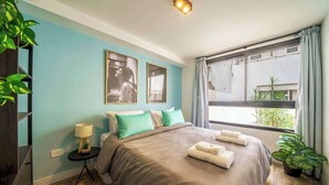 A bedroom with a bed, pillows, a nightstand, a lamp, a window with curtains, and a plant.