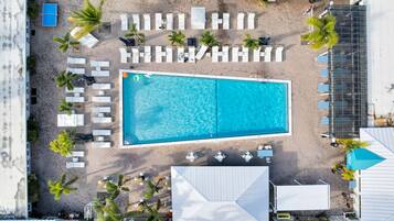 An aerial view of a resort with a large rectangular pool, multiple lounge chairs, and palm trees.