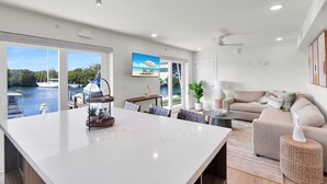 A modern living room with a large white island, a sofa, a coffee table, and a view of boats on the water through large windows. - Waterfront Escape with Pool, Tiki, Breakfast & Marina (Marathon)
