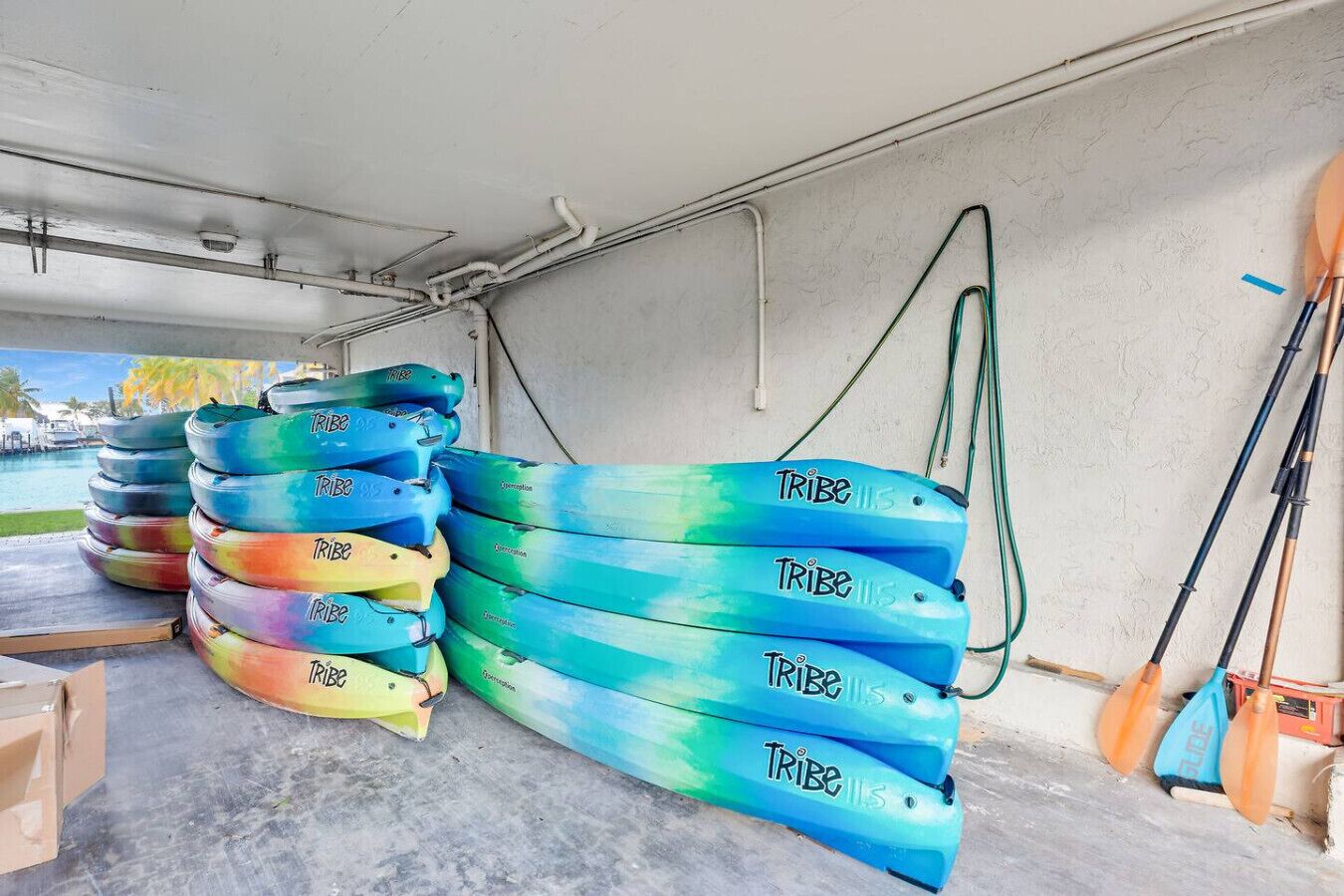 A storage area with several colorful kayaks stacked against a wall, a hose, and a paddle leaning against the wall.