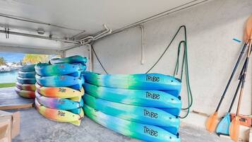 A storage area with several colorful kayaks stacked against a wall, a hose, and a paddle leaning against the wall.