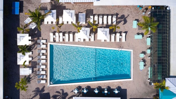 Vista aérea de um resort com uma grande piscina retangular, várias áreas de descanso com cadeiras e guarda-sóis brancos, e palmeiras.
