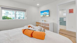 Quarto com cama, janela com vista para edifícios, televisão na parede e banheiro visível por uma porta aberta.