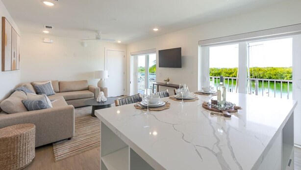 A modern kitchen with a marble countertop, a dining area with a table set for four, and a balcony with a view of greenery. - Boot Key Harbor Retreat with Pool & Breakfast (Marathon)