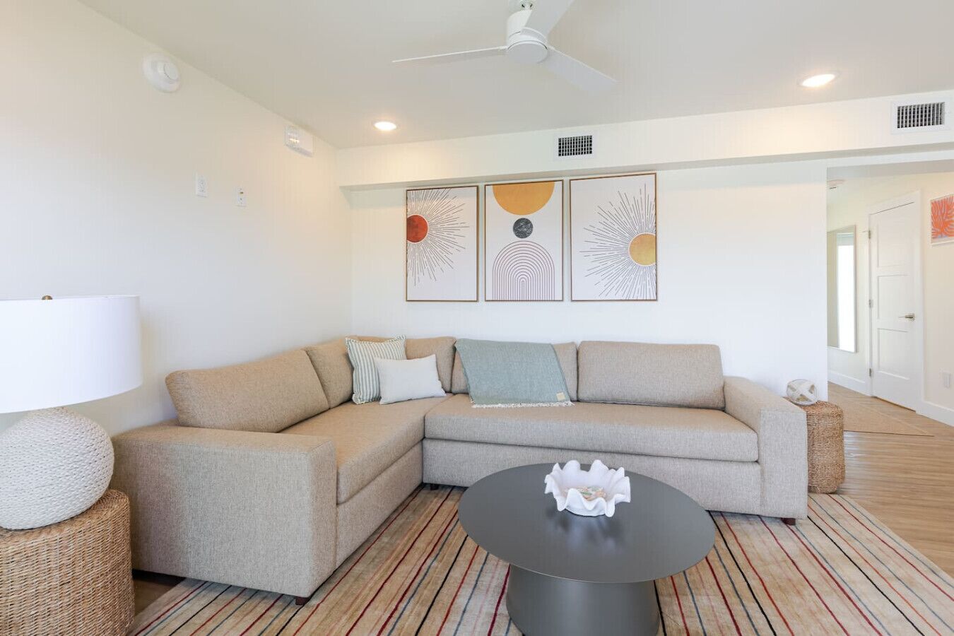 A modern living room with a beige sectional sofa, a round coffee table, and a floor lamp. There are three wall-mounted artworks above the sofa.