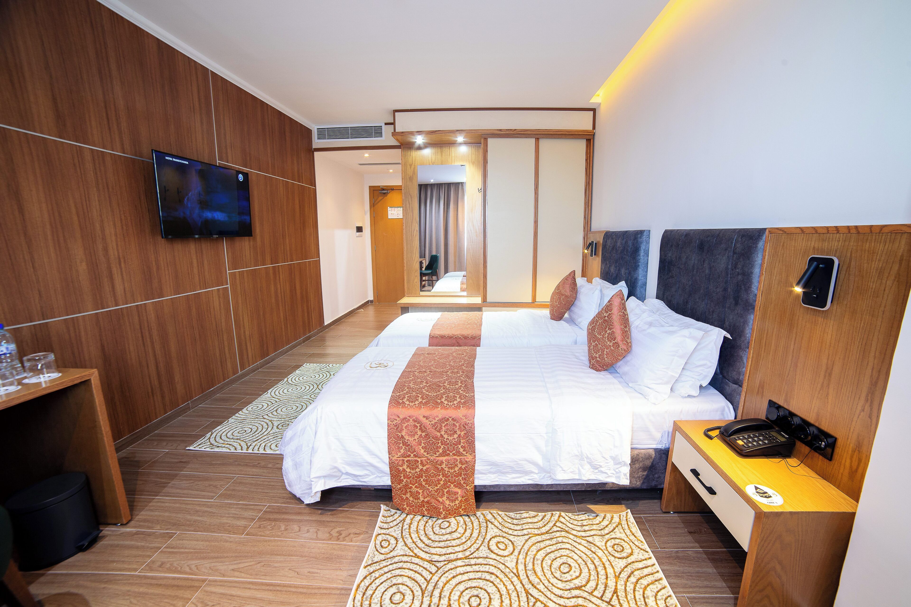 A hotel room with a large bed, a bedside table with a phone, a TV mounted on the wall, and a wooden headboard.