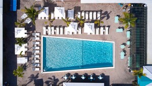 Vue aérienne d’un complexe hôtelier doté d’une grande piscine rectangulaire, entourée de chaises longues blanches et de palmiers.