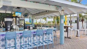 Ein Außenbereich mit Bar, ausgestattet mit einem weißen Sonnendach, blauen Stühlen und einer Theke mit Getränkespender.