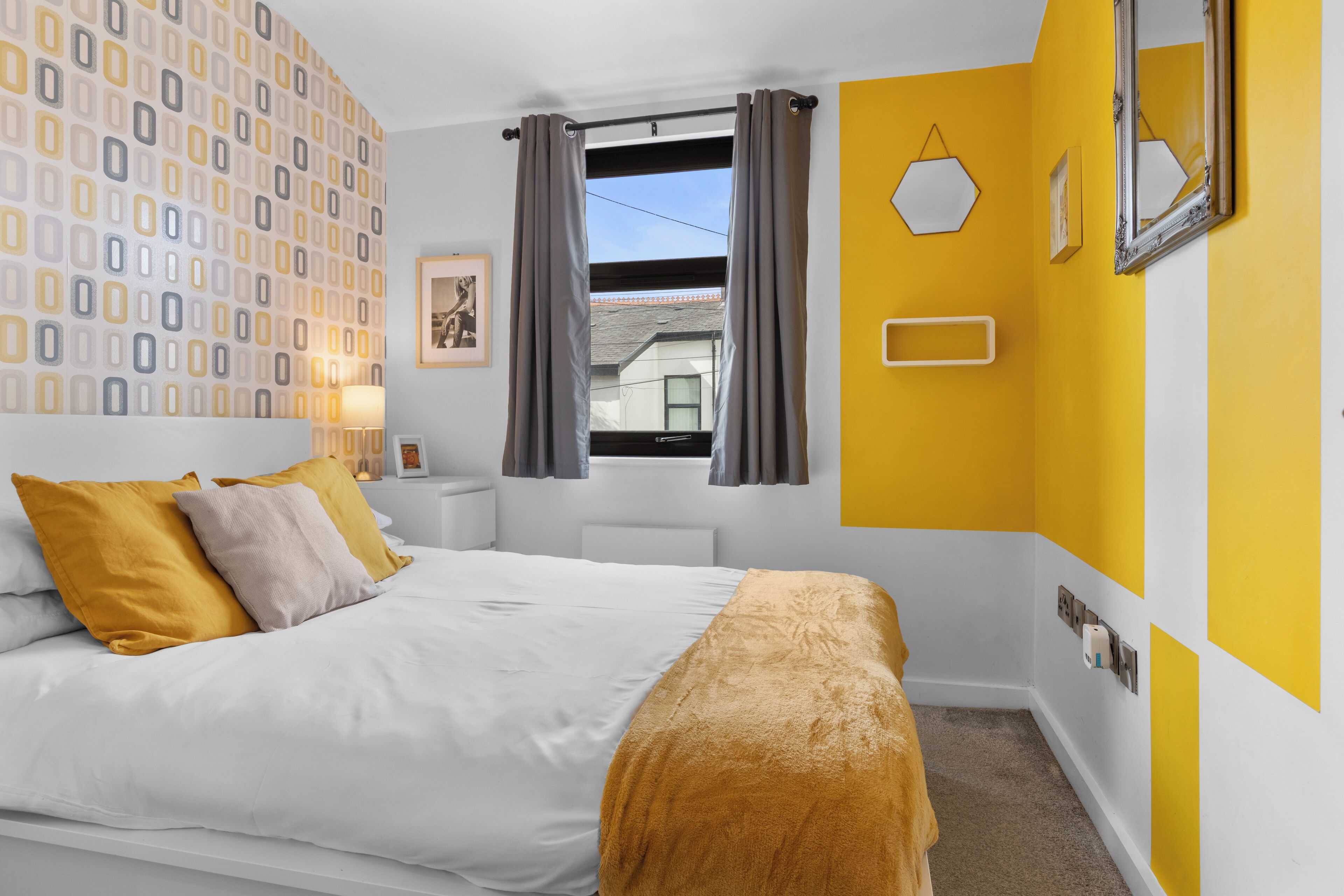 A hotel room with a bed, a window with curtains, a wall with a geometric pattern, and a yellow accent wall.
