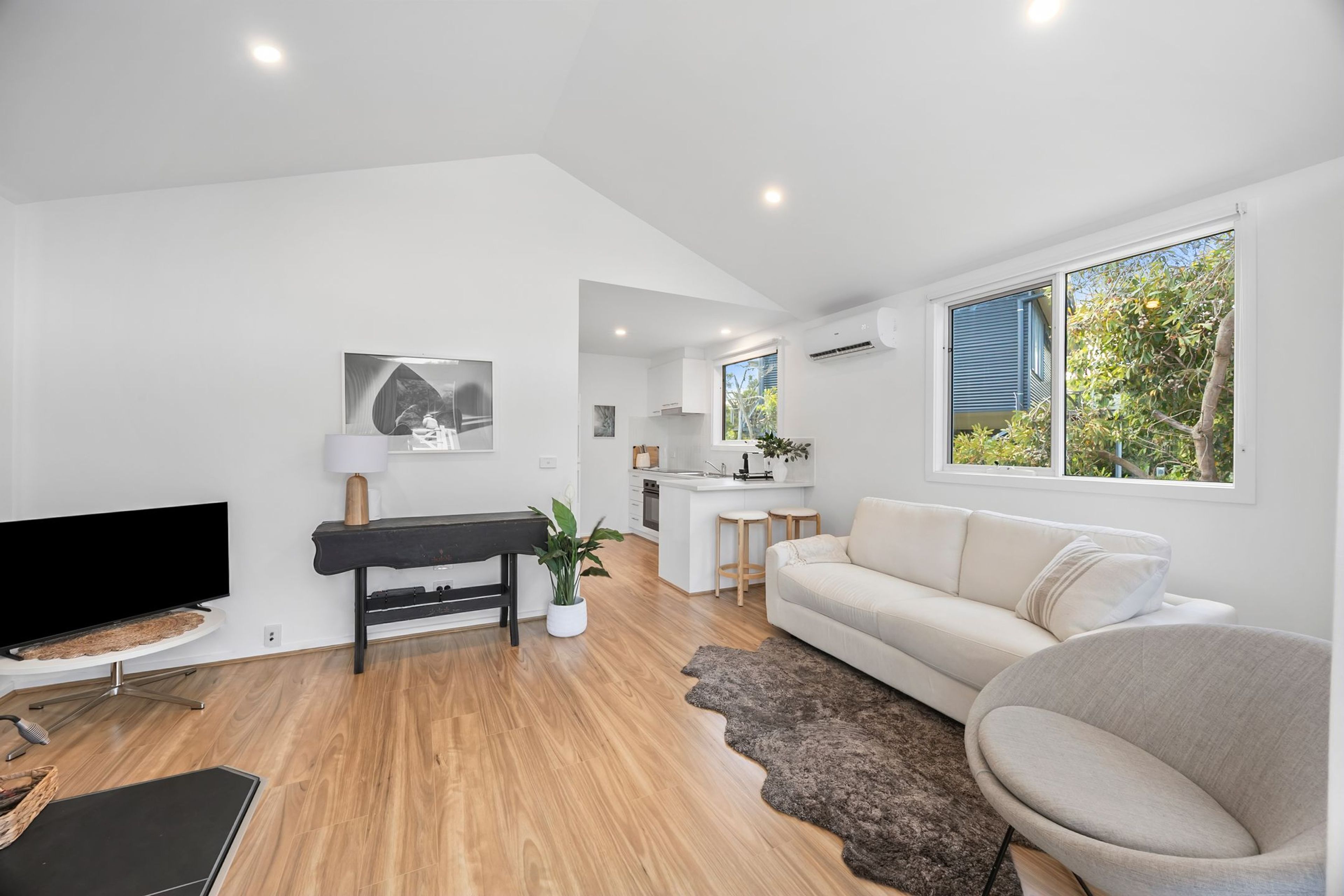 A modern living room with a flat-screen TV, a black bench, a sofa, and a large window with a view of greenery.