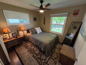 Un dormitorio con cama, dos ventanas, ventilador de techo, mesita de noche, lámpara, espejo y alfombra.