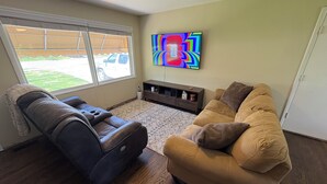 Un salón con un sillón reclinable de cuero, un sofá beige, un televisor con el logotipo de FIFA, un mueble bajo y una ventana con vistas a un coche aparcado y follaje.