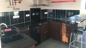 A modern kitchen with black countertops, wooden cabinets, and a stainless steel range hood. There is a built-in dishwasher and a red shelf above the counter. - Gîte Les Rondières (Pougny)