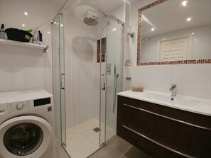 A modern bathroom with a glass-enclosed shower, a built-in washing machine, a vanity with a mirror, and a small shelf with toiletries. - Sea view-Town-House-Comfort-Private Bathroom (Nerja)