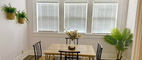 A dining area with a wooden table and four chairs, flanked by three windows with blinds.