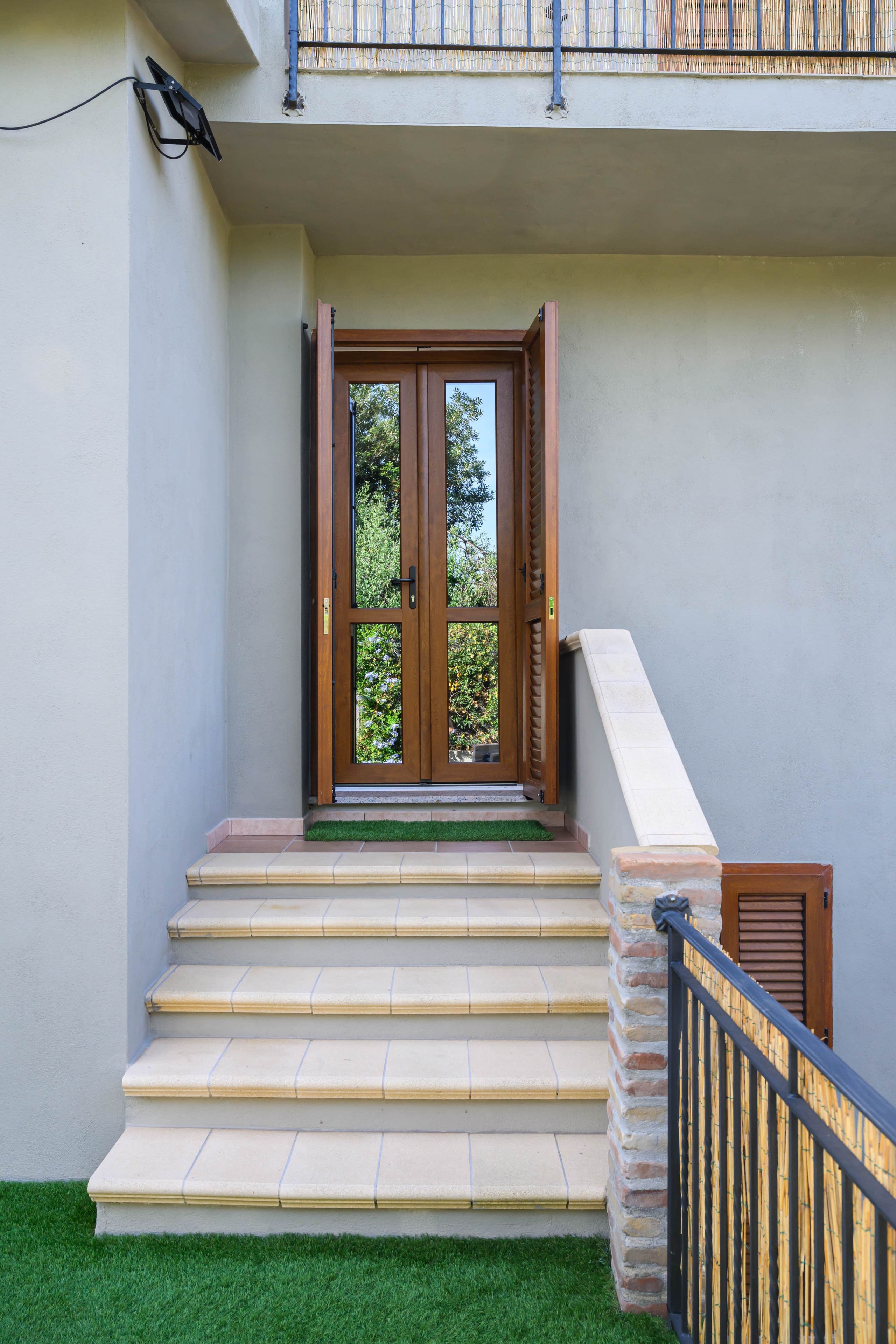 A set of outdoor stairs leading to a wooden door with glass panels, a small garden area with artificial grass, and a metal railing.