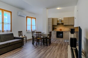 En kompakt opholdsstue med sofa, spisebord, stole, et tekøkken med skabe og hårde hvidevarer, samt et fjernsyn.
