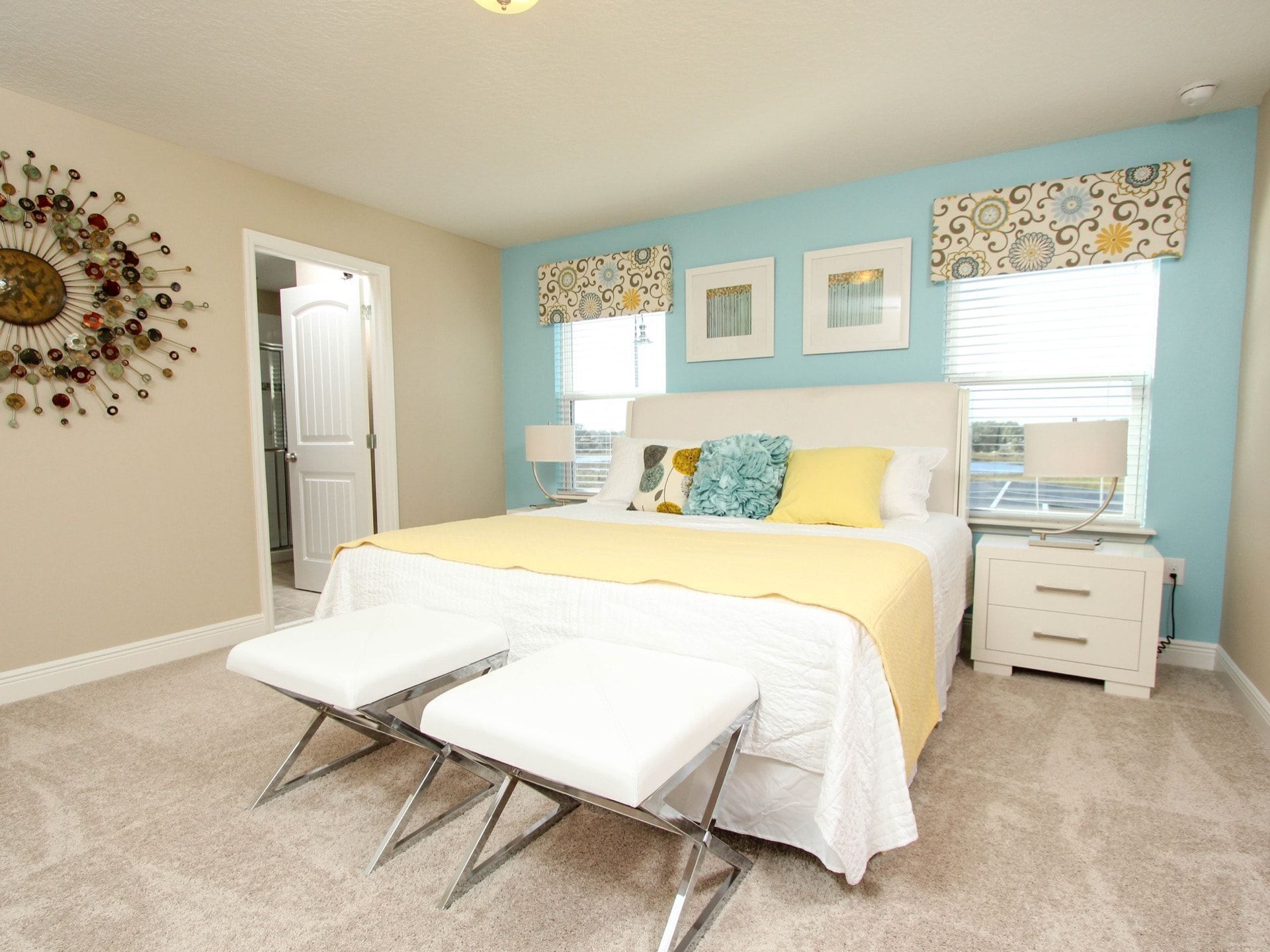 A bedroom with a bed, two white benches, a nightstand, a lamp, and a decorative wall piece.