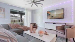 Una sala de estar moderna con un ventilador de techo, un televisor de pantalla plana montado en la pared con una tira de luces LED decorativa, un sofá, una mesa de centro con objetos decorativos y una alfombra estampada.