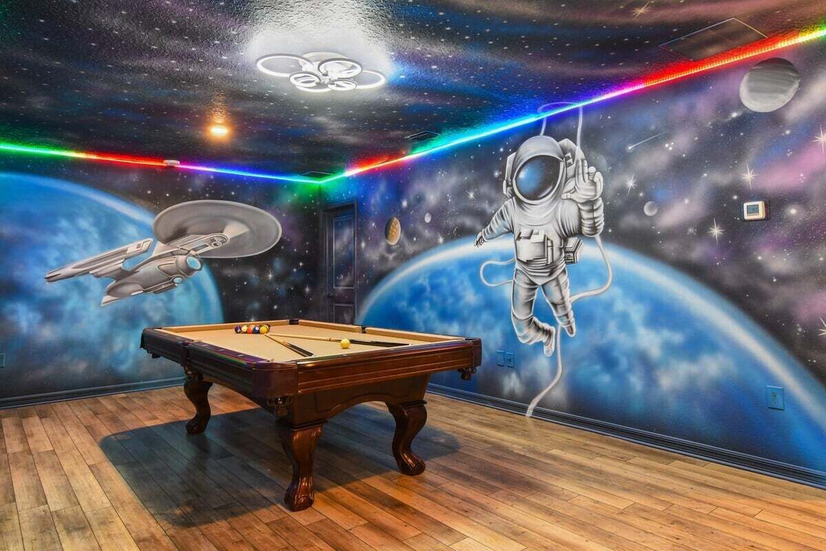 Ein Raum mit Billardtisch, Wandgemälde von Weltraum mit Planeten und Astronaut sowie einer Neonleuchtleiste.