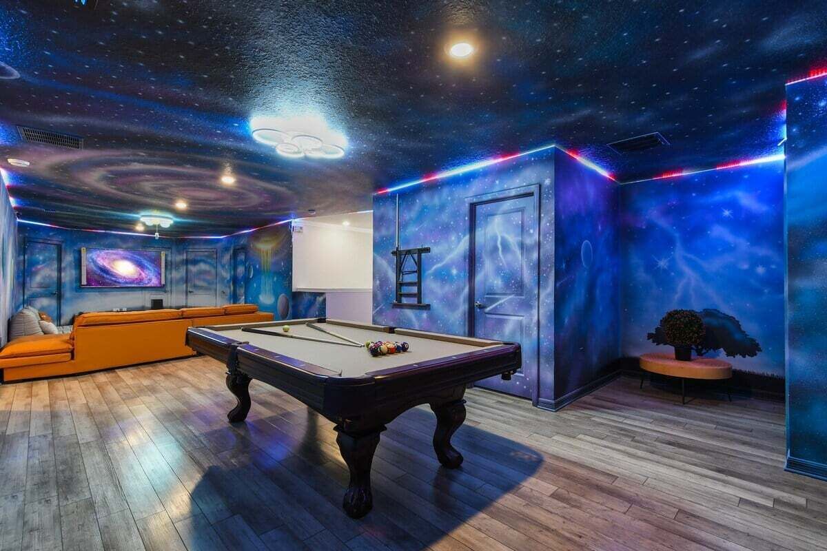 Quarto com mesa de bilhar, sofá e um mural representando uma galáxia.