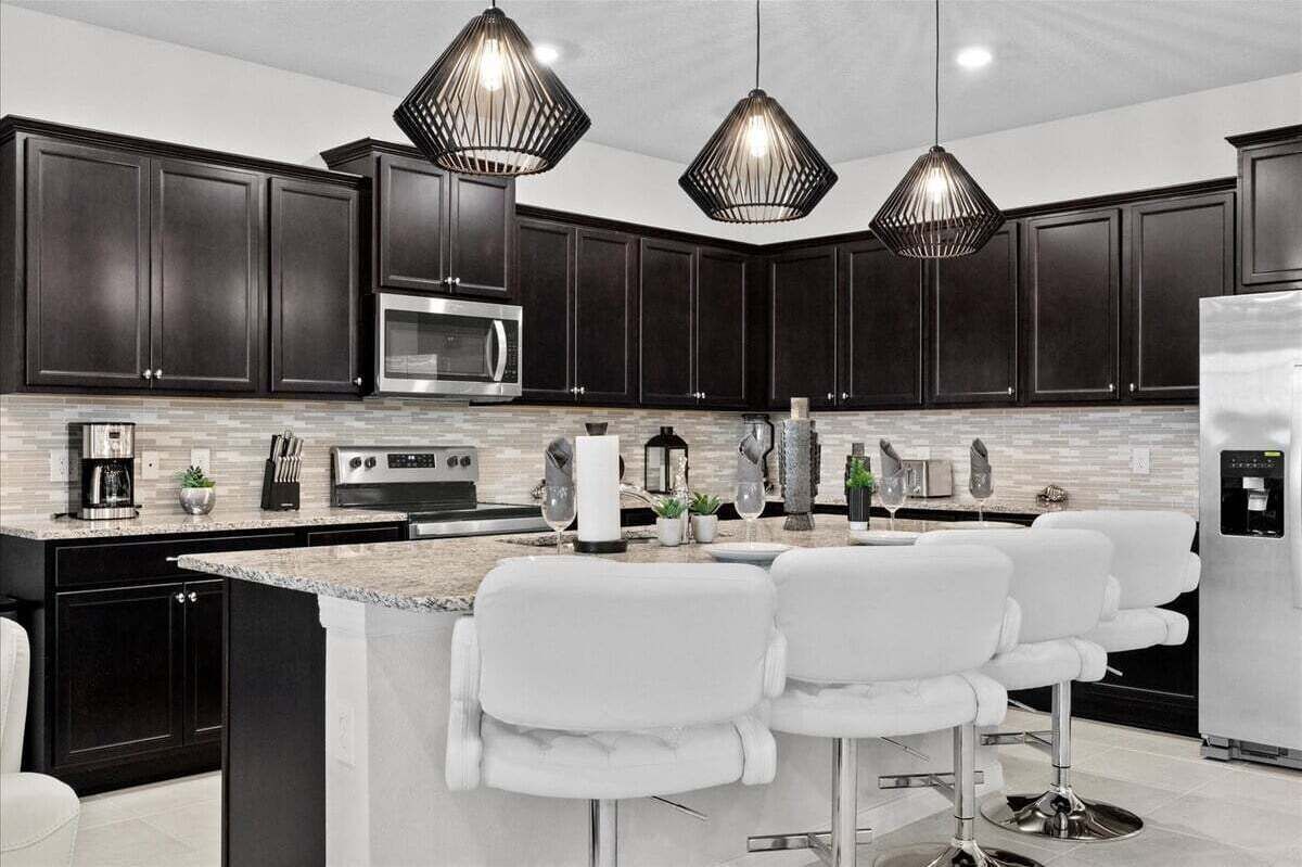 A modern kitchen with dark cabinetry, a central island with bar stools, a stainless steel refrigerator, and pendant lighting.
