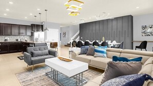 A modern living room with a white coffee table, grey sofa, and a beige sofa with blue cushions. There is a kitchen area with dark cabinets and a dining area with a white table and chairs. - 3739la · Brand New 10br/pool/spa/ Disney Fireworks View (Davenport)