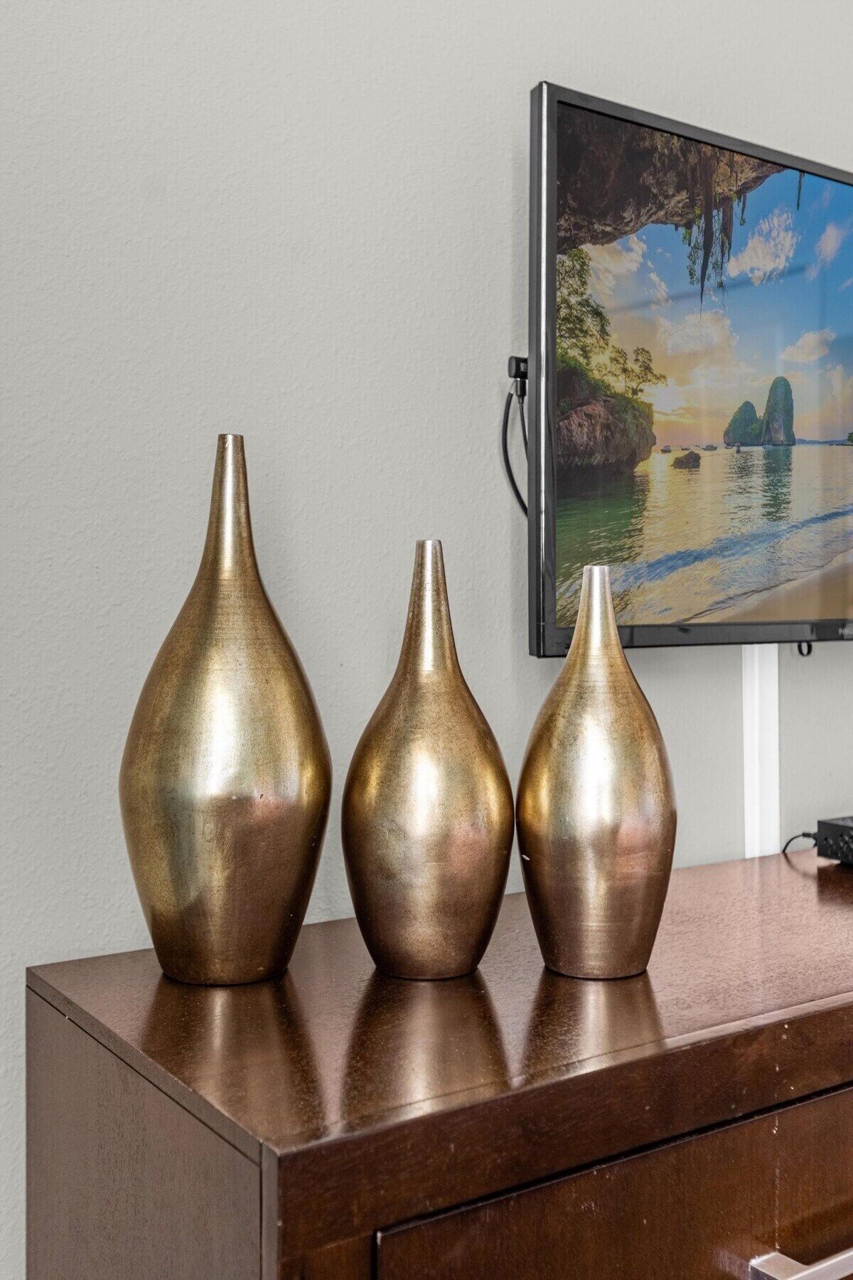 Three metallic vases on a wooden dresser against a wall with a framed picture.
