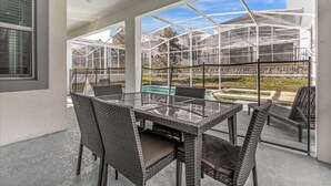 Ein überdachter Patio-Bereich mit einem Glastisch und Korbstühlen, mit Blick auf einen Pool und ein Gewächshaus.