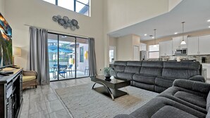 A spacious living room with a grey sectional sofa, a glass coffee table, and a dining area with a table and chairs. A kitchen with white cabinets and modern appliances is visible in the background. - 5424 Solterra Circle, Davenport,5beds (Davenport)