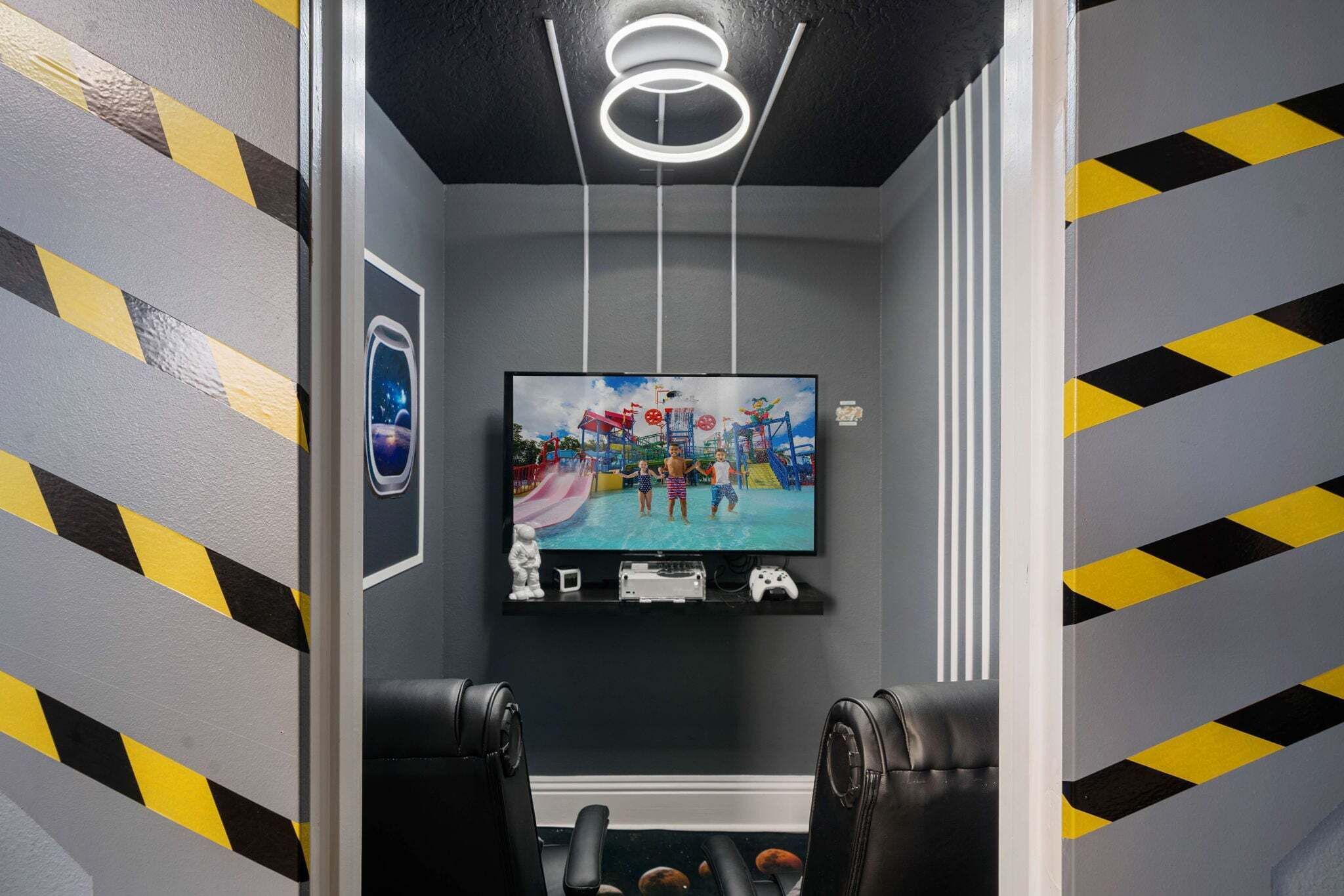 A room with a television mounted on the wall, a gaming console, and two gaming chairs.