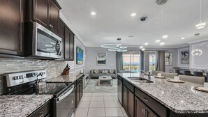 A modern kitchen with dark cabinetry, stainless steel appliances, and a granite countertop. There is a dining area with a sofa and a view of the outdoors. - 3727la - Brand New 10beds Villa at Windsor Island/pool/spa/firew (Davenport)