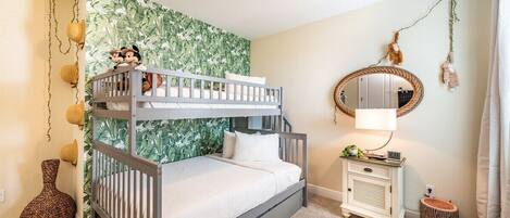 Una cama litera con ropa de cama blanca, una mesita de noche pequeña con una lámpara, un espejo y una pared decorativa con un diseño de hojas verdes.