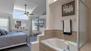 A bedroom with a large bed, a ceiling fan, a vanity with a mirror, and a bathroom with a bathtub and a shower. - 15730 BAY Vista Drive Clermont, 4bed (Clermont)