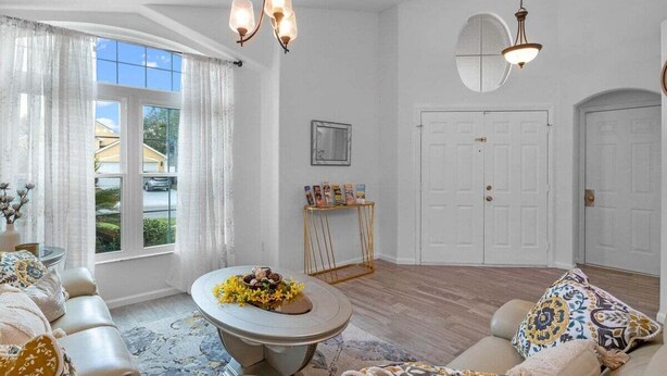 A spacious living room with a large window, a round coffee table with flowers, a sofa, and a decorative mirror. - 15730 BAY Vista Drive Clermont, 4bed (Clermont)