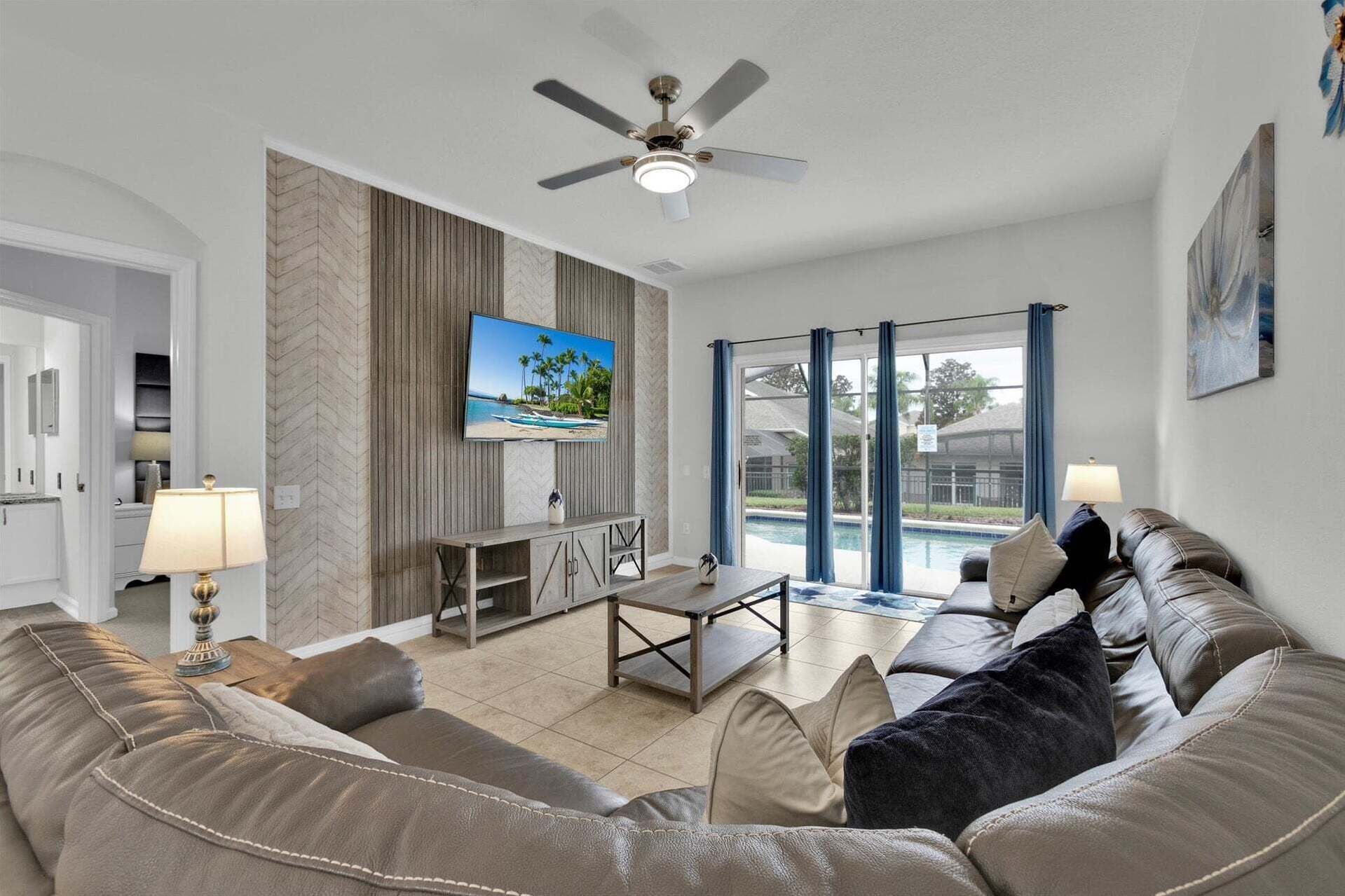 A living room with a sofa, a TV, a ceiling fan, and a view of a swimming pool.