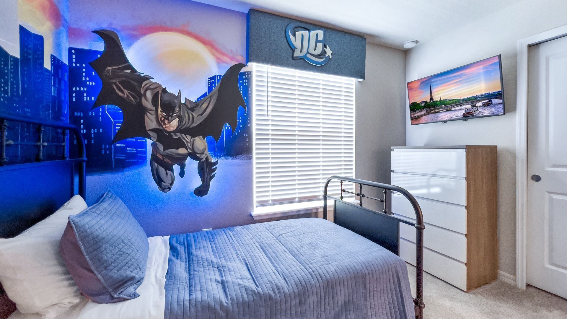 Une chambre avec une fresque représentant un paysage urbain, un lit recouvert d’une couette grise, une fenêtre avec des stores, une commode blanche et un tableau représentant la Tour Eiffel.