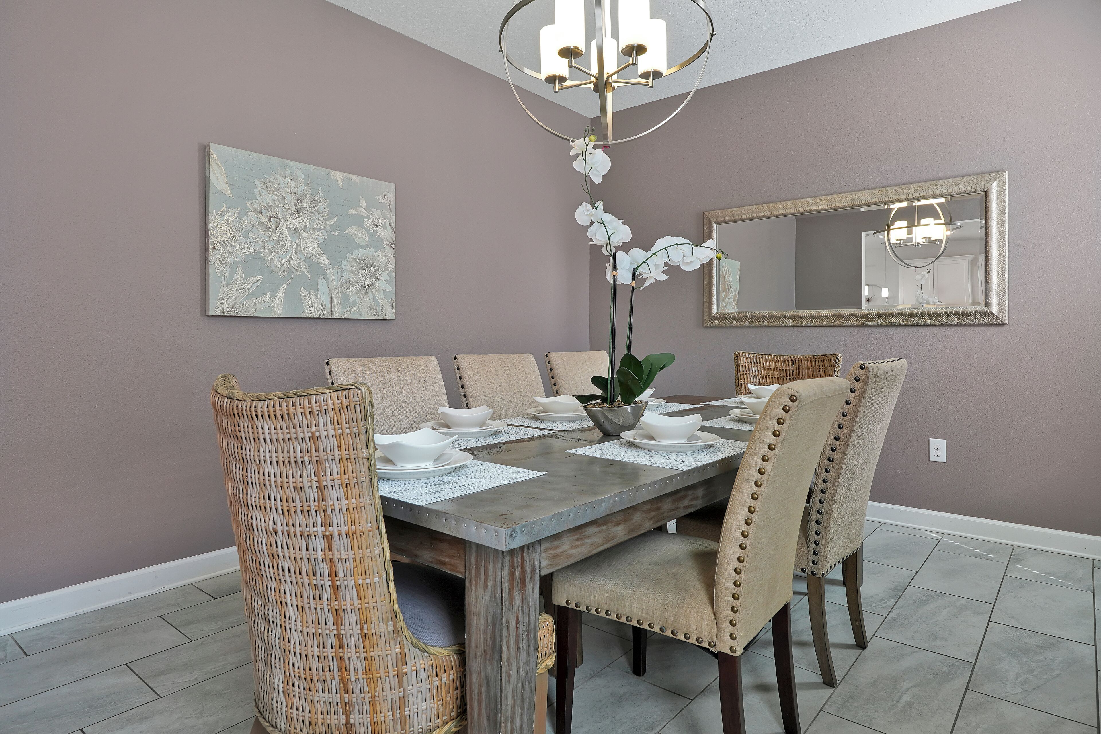 A dining area with a wooden table, beige upholstered chairs, a woven chair, a potted orchid, and a painting on the wall.