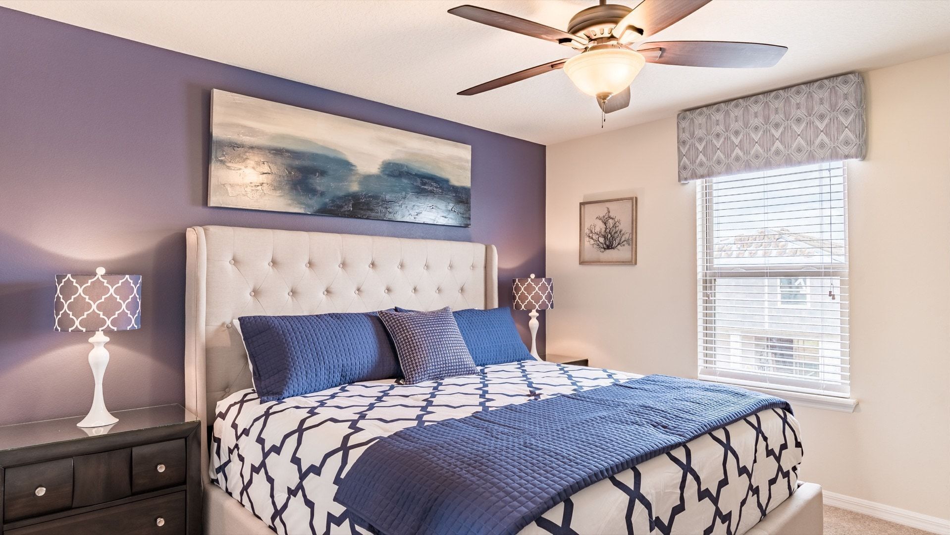 A bedroom with a tufted headboard, a large bed with patterned bedding, a ceiling fan, a window with blinds, and a wall-mounted abstract painting.